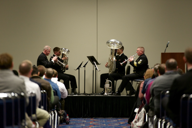 Eine Gruppe von Männern, die auf einer Bühne Blasinstrumente spielen, mit Notenständern, einem Podium und einem Mikrofon, vor einem Publikum, das auf Stühlen sitzt.