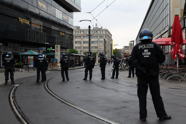 Eine Gruppe von Polizeibeamten in schwarzen Uniformen und Helmen steht auf einer Straße, die von Glasfenster-Gebäuden gesäumt ist, mit Bäumen, Strommasten und einem klaren Himmel im Hintergrund, während Schirme, Stühle und Tische in der Nähe verstreut sind.