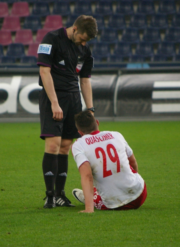 Ein Fußballspieler in Sportbekleidung sitzt neben einem Schiedsrichter auf dem Boden, beide in einer Stadionumgebung mit Tafeln und Stühlen im Hintergrund.