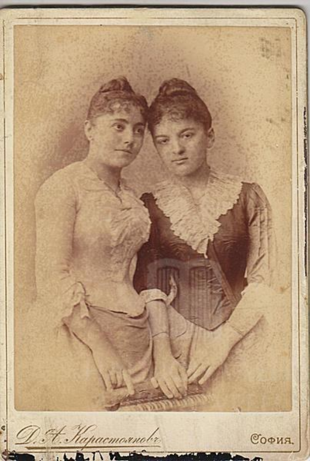 Zwei junge Frauen in traditioneller russischer Kleidung posieren gemeinsam auf einem alten Foto, eine in einem weißen Kleid und die andere in einem blauen Kleid, vor einem hellblauen Hintergrund mit Text unten.