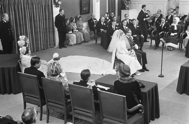 Schwarzes und weißes Foto einer Hochzeitszeremonie mit Sitzenden und Stehenden, einem Mann mit Mikrofon und einer Frau mit Blumenstrauß, sowie einem Podium und Blumenschmuck.