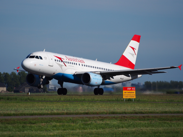 Airbus A320-200 der Austrian Airlines startet vom Frankfurter Flughafen mit einer Tafel im Vordergrund, Gras, Bäumen, Gebäuden und einem klaren blauen Himmel im Hintergrund.