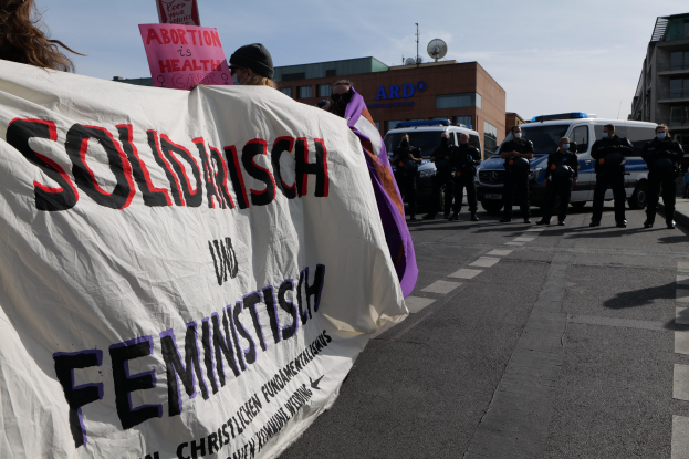 Eine Gruppe von Menschen marschiert auf der Straße, trägt eine Fahne mit der Aufschrift 'Solidarität und Feminismus', mit geparkten Fahrzeugen und Gebäuden im Hintergrund unter einem klaren blauen Himmel.