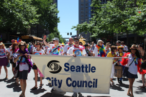 Eine Gruppe von Menschen marschiert mit einem "Seattle City Council"-Schild die Straße entlang, einige tragen Mützen und Schutzbrillen, andere tragen Taschen, mit Bäumen, Gebäuden und einem klaren blauen Himmel im Hintergrund.