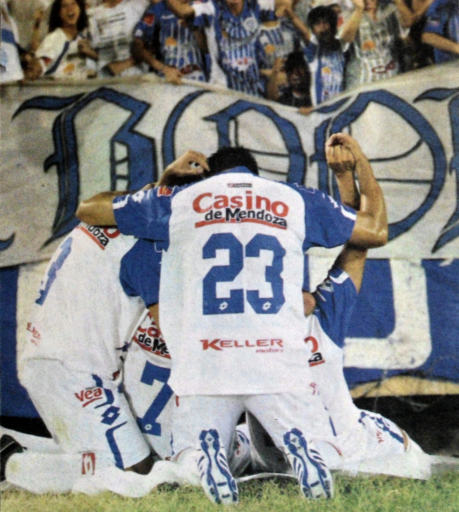 Gruppe von Fußballspielern feiert vor jubelnder Menge, mit einem Banner im Hintergrund.