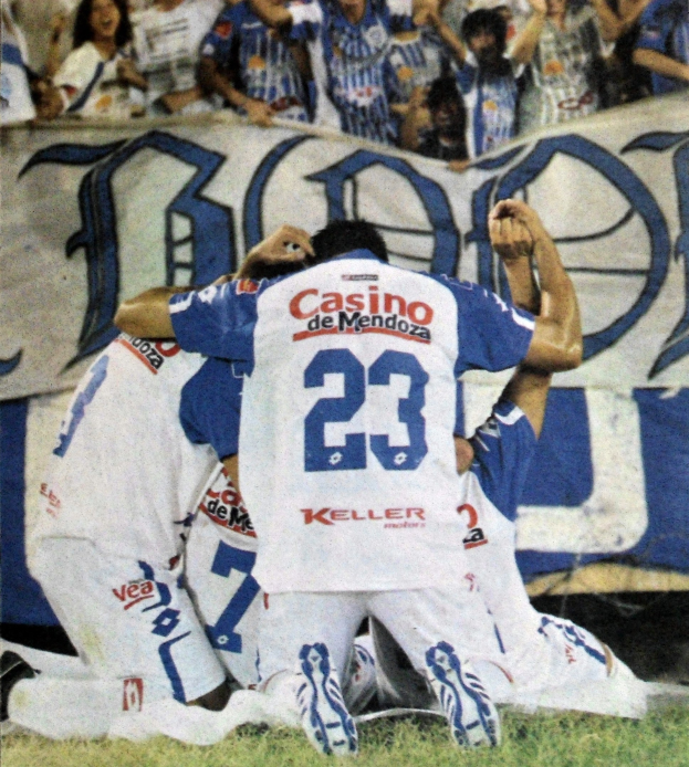 Gruppe von Fußballspielern feiert vor jubelnder Menge, mit einem Banner im Hintergrund.
