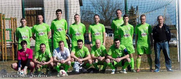 Eine Gruppe von Männern in grünen T-Shirts steht auf einem Fußballfeld in der Nähe eines Torpfostens, mit einem Ball davor, während Bäume, Gebäude und Fahrzeuge im Hintergrund zu sehen sind.
