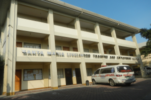 Außenansicht des Gebäudes der Santa Maria Lebensmittelausbildung und Beschäftigungszentrum mit Fenstern, Türen und Beschriftung, umgeben von Bäumen mit Fahrzeugen auf der Straße und einem klaren blauen Himmel.