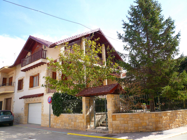 Eine Villa mit einem vor ihr geparkten Auto, mit Fenstern, Geländern, Pfeilern und einem Dach, umgeben von einem Tor, einem Schild, einem Straßenpfahl, Bäumen und einer Wand unter einem bewölktem Himmel.