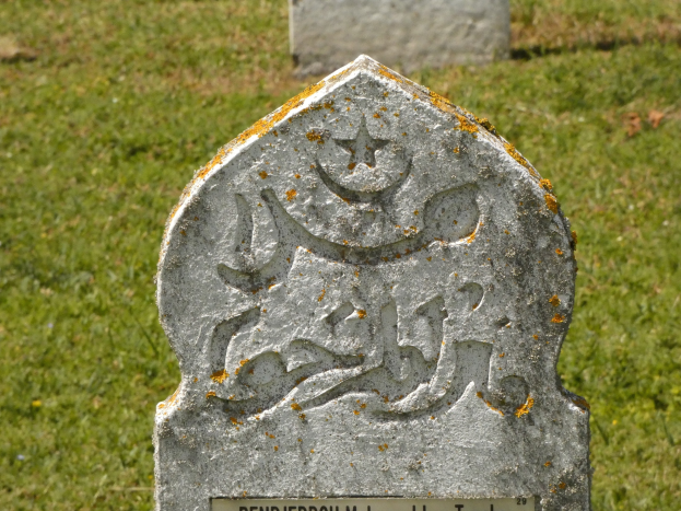 Grabstein mit arabischer Inschrift in einem grünen Friedhof.