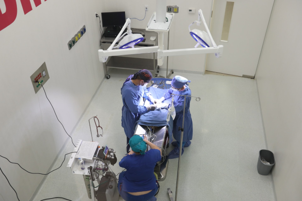 Eine Gruppe von Chirurgen führt einen Eingriff in einem Operationssaal durch, wobei eine Person auf einem Operationstisch umgeben von medizinischer Ausrüstung liegt, eine beschriftete Wand links, ein Objekt auf dem Boden rechts und verschiedene medizinische Geräte im Hintergrund.