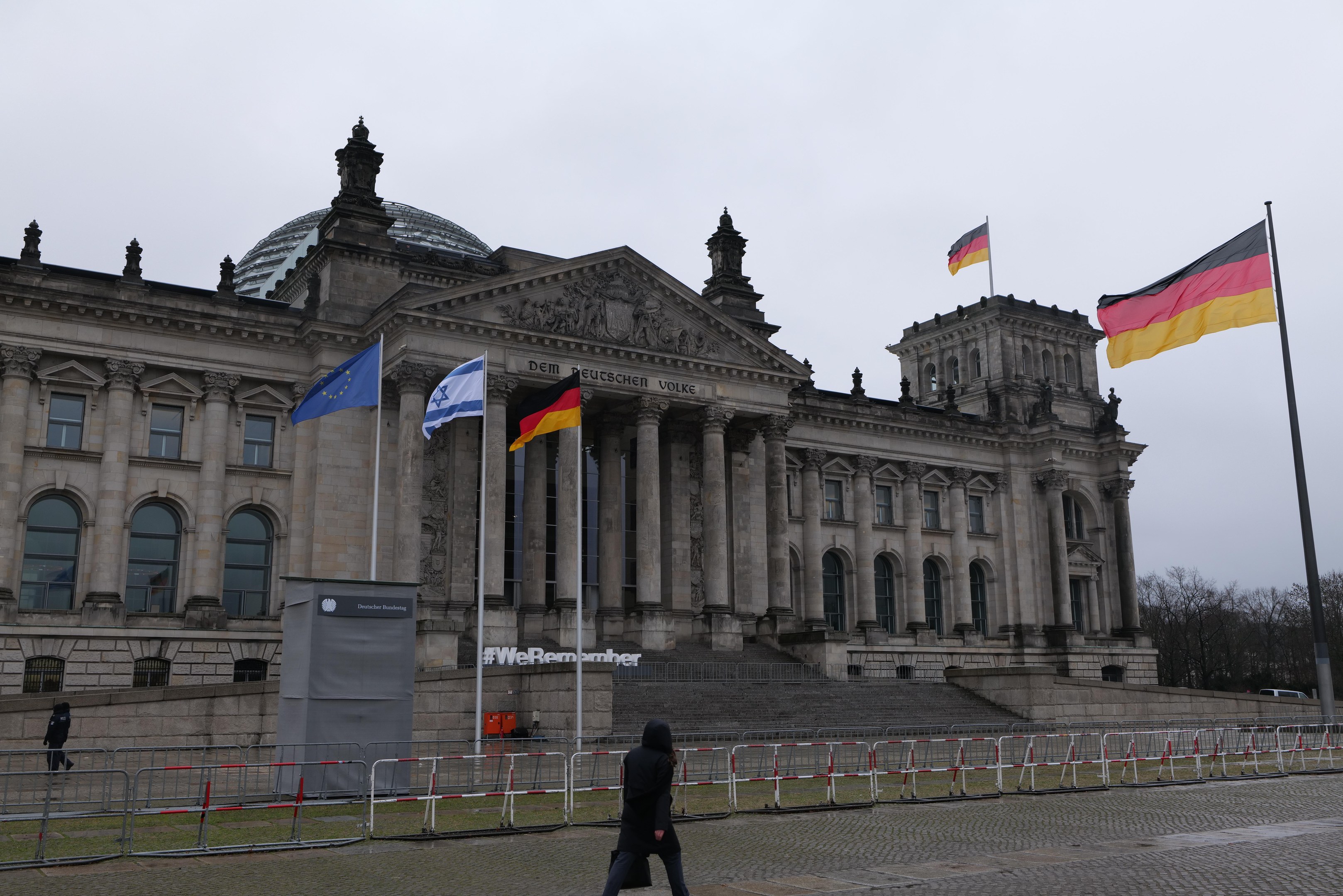 Eine Person, die vor dem Reichstagsgebäude in Berlin, Deutschland, mit seinen Fenstern, Säulen, Bögen und Statuen, umgeben von Fahnenmasten, Treppen, Geländern, Bäumen und einem klaren blauen Himmel, spaziert.