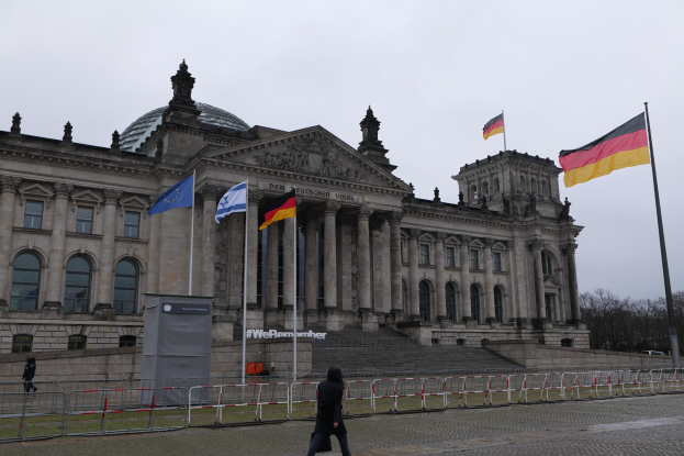 Eine Person, die vor dem Reichstagsgebäude in Berlin, Deutschland, mit seinen Fenstern, Säulen, Bögen und Statuen, umgeben von Fahnenmasten, Treppen, Geländern, Bäumen und einem klaren blauen Himmel, spaziert.