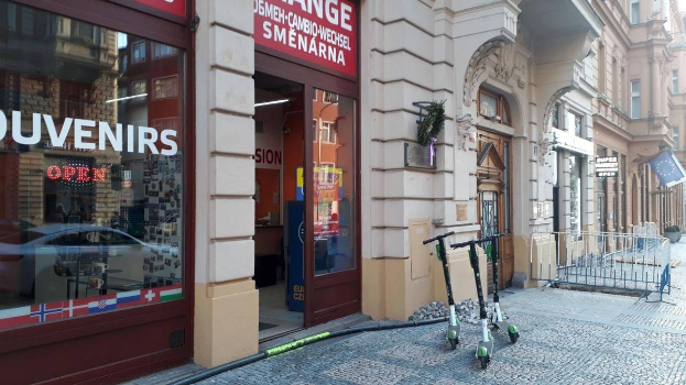 Eine Straße in einer Stadt mit parkenden Rollern, Gebäuden, Informationsschildern, einem Geländer, Bäumen und einem klaren blauen Himmel.