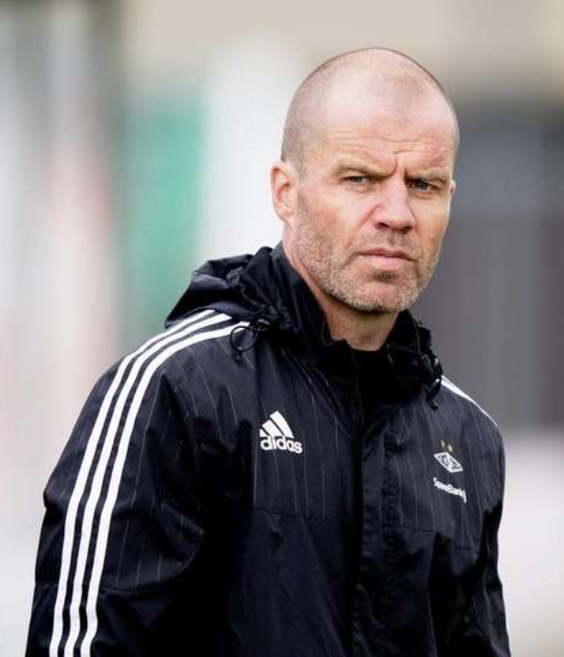 Ein kahlgeschorener Trainer in einer blauen Jacke mit weißen Streifen und einem weißen Kragen steht auf einem Fussballfeld, die Hände verschränkt und einen ernsten Ausdruck, mit einem unscharfen Hintergrund.