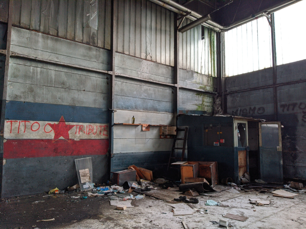 Verlassener Gebäuderaum mit Schutt, einschließlich Kartons, einer Leiter und verstreuten Gegenständen, eine Wand mit Text auf der linken Seite und Glasfenster auf der rechten Seite.