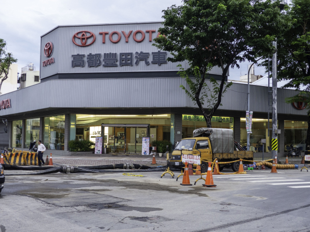 Außenansicht eines Toyota-Autohauses in Tokio, Japan, mit Fahrzeugen, Verkehrskegeln, Rohren, Fußgängern, Bäumen, Strommasten, Schildern und umliegenden Gebäuden unter einem bewölkten Himmel.