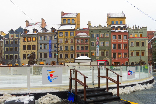 Eisbahn in einem Stadtplatz umgeben von Gebäuden, Treppen, Geländern, Informationsschildern, schneebedeckter Erde, Laternen, Bäumen und einem Himmel als Hintergrund.