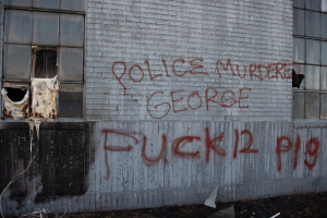 Ein Gebäude mit Graffiti, das "Polizei ermordet George Fuck 12 P19" auf der Seite trägt, mit Fenstern und verschiedenen Gegenständen vorne.