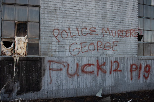 Ein Gebäude mit Graffiti, das "Polizei ermordet George Fuck 12 P19" auf der Seite trägt, mit Fenstern und verschiedenen Gegenständen vorne.