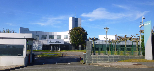 Ein großes, mehrfenstriges Gebäude mit einem Tor, umgeben von Straßenmasten, Laternen, Gittern, Bäumen und Gras, unter einem bewölkten Himmel, das als Hauptsitz von Iberostica in den Niederlanden dient.