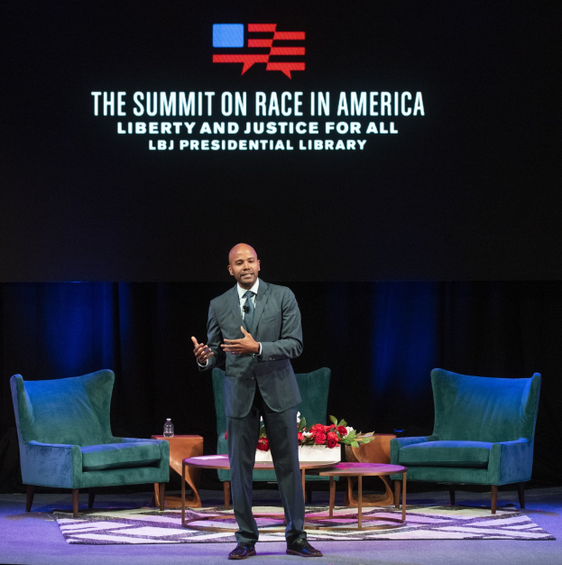 Ein Mann in einem Blazer, Krawatte und Hemd steht auf einer Bühne und spricht bei der "The Summit on Race in America: Liberty and Justice for All"-Veranstaltung in der LBJ Presidential Library, mit zwei Stühlen, einem Tisch mit einem Blumenstrauß und einer Flasche darauf und einem Bildschirm dahinter.