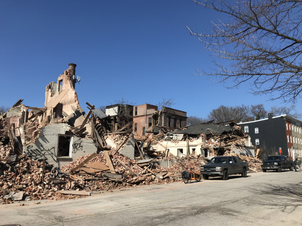 Ein eingestürztes Gebäude an einer Straßenecke mit verstreuter Trümmern, Fahrzeugen auf der Straße, Passanten auf dem Gehweg, Bäumen und einem klaren Himmel.