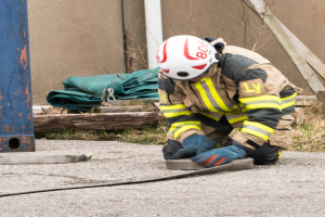Ein Feuerwehrmann in Schutzausrüstung kniet auf dem Boden und verwendet einen Schlauch, um ein Feuer zu löschen, wobei verschiedene Gegenstände und ein metallisches Objekt im Hintergrund zu sehen sind.