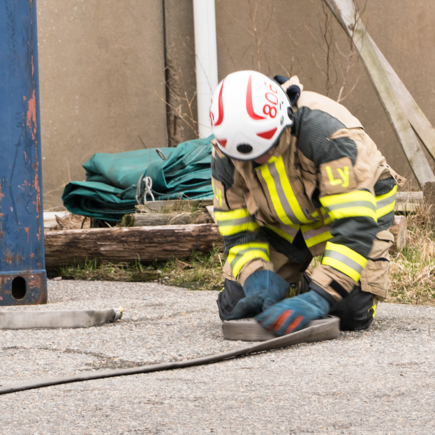 Ein Feuerwehrmann in Schutzausrüstung kniet auf dem Boden und verwendet einen Schlauch, um ein Feuer zu löschen, wobei verschiedene Gegenstände und ein metallisches Objekt im Hintergrund zu sehen sind.