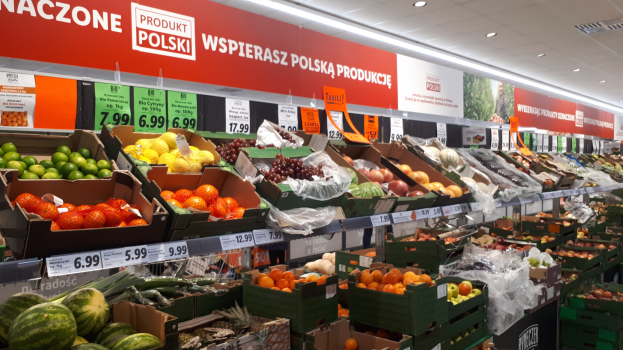 Ladeninneres mit verschiedenen Obst- und Gemüsesorten in Kartonschachteln mit Preisschildern, Wandtafeln mit Text und Bildern sowie Deckenbeleuchtung.