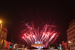 Eine belebte Stadtstraße während einer Silvesterfeier in Berlin, voller Menschen, Fahrzeuge und festlicher Lichter von Gebäuden und Feuerwerk am Himmel.