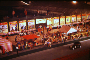 Ein altes Nachtumzugsbild mit Menschen, einem Auto, Masten mit Fahnen, einem Zaun, einem Zelt, Brettern mit Bildern und Text sowie einer Straßenlaterne.