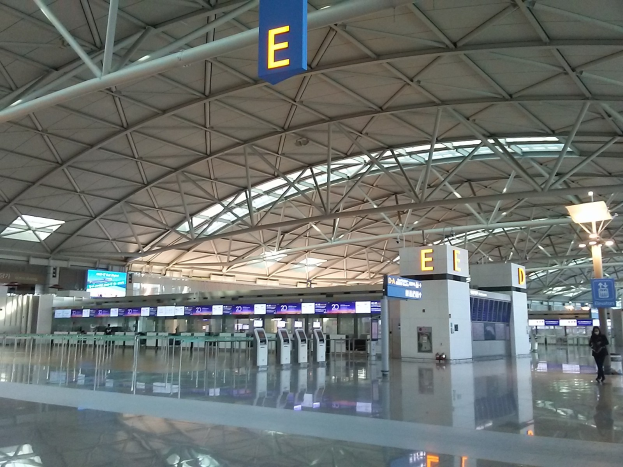 Leerer Flughafen-Terminal mit Menschen, die gehen, Schilder mit Text und Maschinen in der Mitte, eine Person links und Metallstangen und Lampen an der Decke.