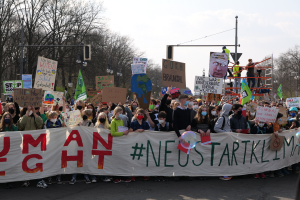 Eine große Gruppe von Menschen marschiert auf einer Straße, hält eine 'Menschliche Rechte'-Schriftzug-Tafel und verschiedene Plakate, mit Bäumen, Laternenmasten und einem klaren blauen Himmel im Hintergrund.
