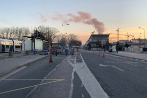 Eine Straßenansicht mit Verkehrskegeln, einem Bus, Laternenmasten, Bäumen, Gebäuden, Kränen und Rauch, der aus einem entfernten Gebäude aufsteigt, unter einem klaren Himmel.