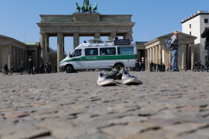 Polizeifahrzeug vor dem Brandenburger Tor mit Menschen und Fahrrädern in der Nähe, ein Paar Schuhe auf dem Boden und der Bogen mit Statuen, Bäumen und einem klaren Himmel im Hintergrund.