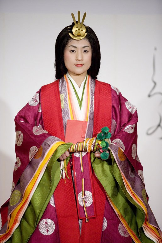 Eine Frau in einem leuchtend roten und grünen traditionellen japanischen Kimono mit aufwändigen Mustern steht selbstbewusst auf einem Laufsteg, ihr Haar ist zu einem ordentlichen Knoten gebunden.