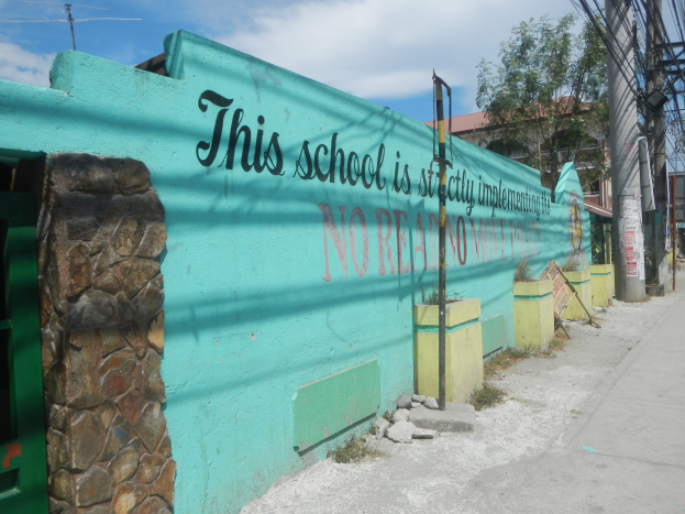 Grüne Wand mit Graffiti 'Diese Schule ist keine Stadt' umgeben von Gebäuden, Bäumen, Strommasten, Drähten und anderen städtischen Elementen unter einem bewölkten Himmel.