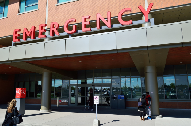 Gruppe von Menschen vor einem Notaufnahmesgebäude mit Glaswänden, Säulen und Beschriftung.