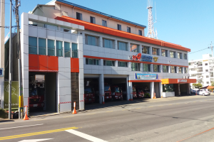 Eine Feuerwache mit einem Feuerwehrauto davor, umgeben von Gebäuden, Fahrzeugen, Verkehrskegeln, Strommasten, Kabeln, Namensschildern, Bäumen und einem klaren blauen Himmel.