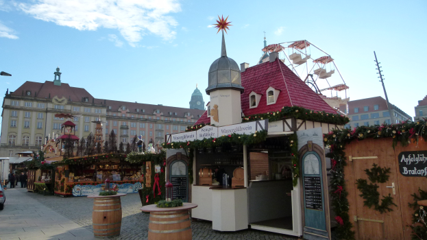Ein geschäftiger Weihnachtsmarkt auf einem Stadtplatz mit geschmückten Bäumen, Gebäuden, Straßenlaternen, Fahrzeugen, Passanten und einem bewölkten Himmel.