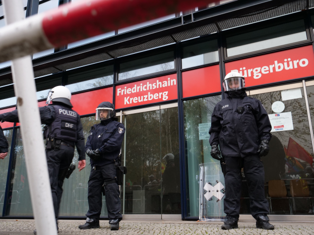 Polizeibeamte in Helmen und Handschuhen stehen vor einem Gebäude mit Glaswänden und -türen, in der Nähe eines Pfahls und Schildern mit der Aufschrift "Friedrichshain Kreuzberg".
