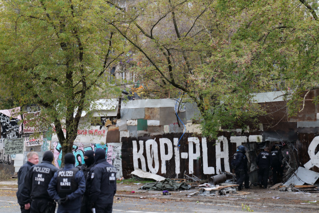 Eine Gruppe von Polizisten in schwarzen Uniformen und Helmen steht vor einem mit Graffiti bedeckten, baufälligen Gebäude mit verstreuter Trümmern und Bäumen im Hintergrund.