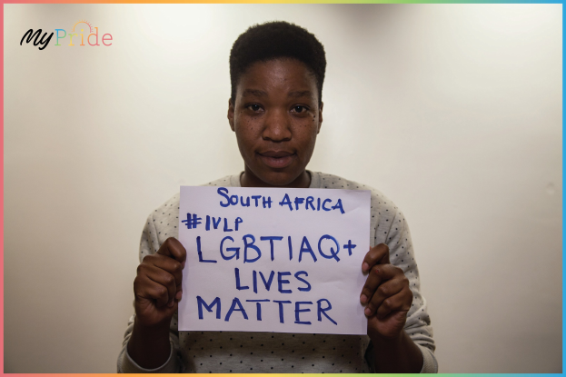 Eine Frau in einem weißen Hemd steht entschlossen vor einer Wand und hält ein Schild mit der Aufschrift "LGBTQ+ Lives Matter" in fetten schwarzen Buchstaben hoch.