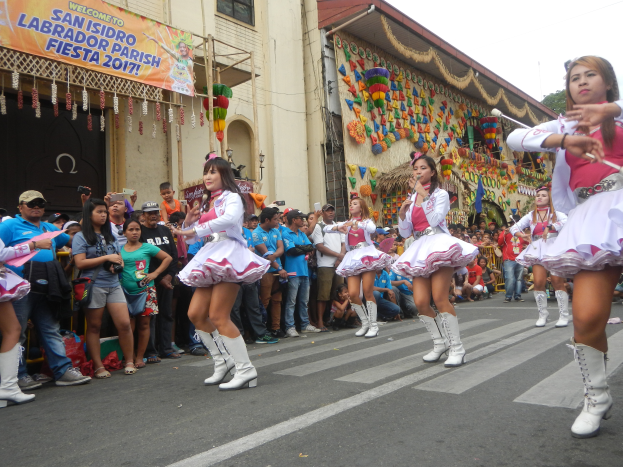 Eine Gruppe von Frauen in pinken und weißen Outfits tanzt auf der Straße, umgeben von einer Menge Menschen, die stehen und zuschauen, mit Gebäuden, Bäumen und einem klaren blauen Himmel im Hintergrund.