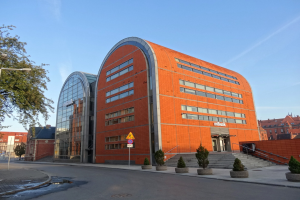 Ein großes oranges IBM-Hauptquartiergebäude mit vielen Fenstern, umgeben von Straßenelementen, Fahrzeugen und Topfpflanzen unter einem bewölkten Himmel.