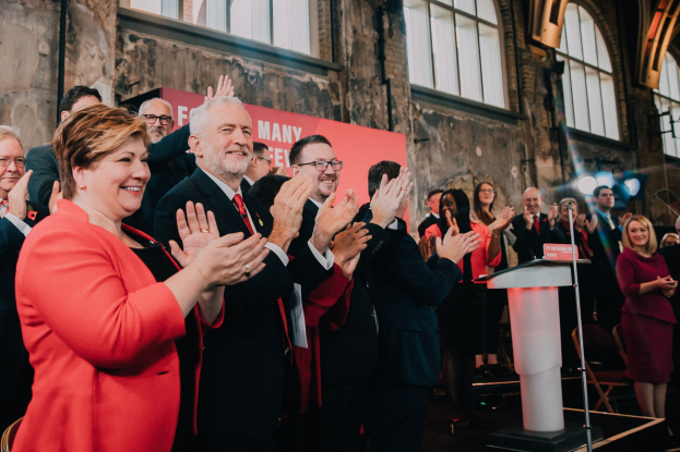 Eine Gruppe von Menschen vor einem Publikum, das klatscht, mit einem Podium, einem Mikrofon und einer Tafel mit Text auf der rechten Seite sowie Stühlen, einer Fahne, einer Wand, Fenstern und Lichtern im Hintergrund.