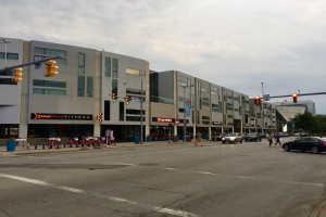 Eine Stadtstraßenszene mit parkenden Autos, Fußgängern, Fahrrädern, Ampeln, Straßenlaternen, Schildern, Bäumen und Gebäuden, mit einem orangefarbenen Fitnessstudio in der Mitte.