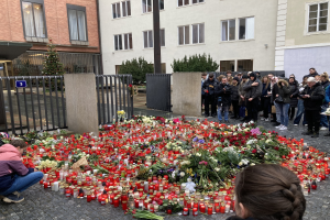 Eine Gruppe von Menschen an einer improvisierten Gedenkstätte mit Blumen, Kerzen und Gegenständen vor einem Gebäude, eine Person kniend mit einer Kamera, mit Gebäuden, Pflanzen, einem Pfahl und einem Tor im Hintergrund.