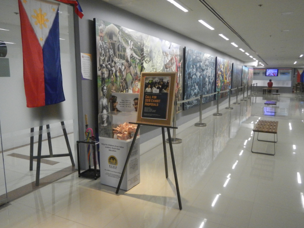 Langer Flur mit einer philippinischen Flagge an der Wand, Displaytafeln mit Text und Bildern, Ribbonständer, Bänke, Deckenleuchten und ein Bildschirm im Hintergrund mit Museumsinformationen.